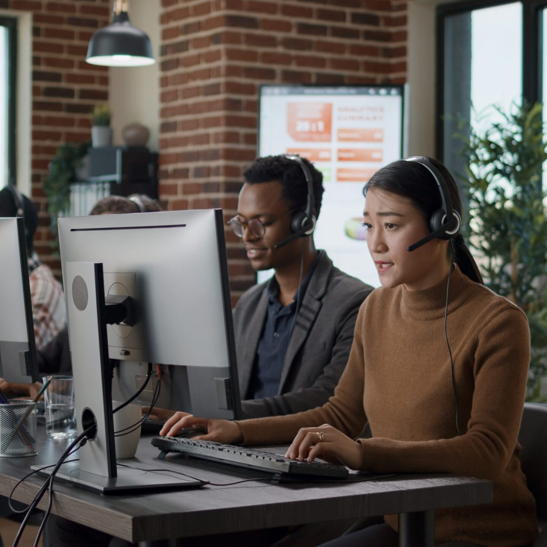 Multiethnic team of people working at call center office, using audio headset for telecommunications to help clients. People answering call on phone helpline, giving assistance at workstation.